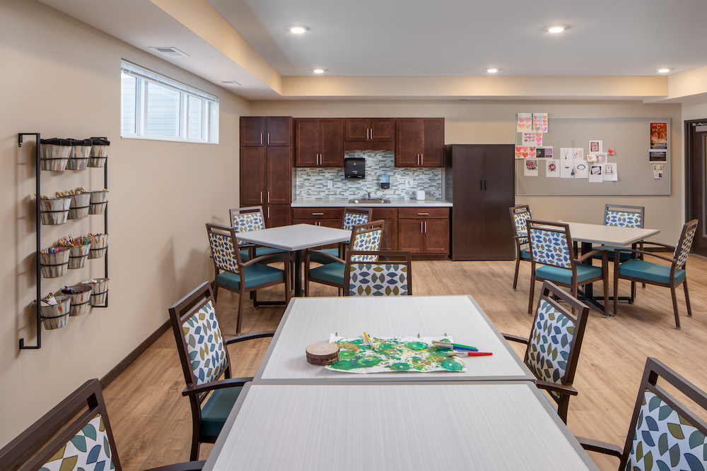 Bright activity room with tables, chairs, and art supplies, featuring large windows and wooden cabinets.