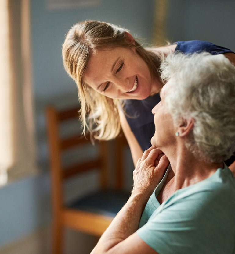 _-senior-memory-care-caregiver- Caregiver smiling warmly at a senior woman in a cozy room during a caregiving session.