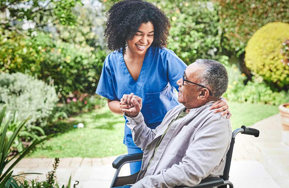 senior-memory-care-caregiver- Caregiver in blue scrubs smiling at a senior man in a wheelchair in an outdoor garden