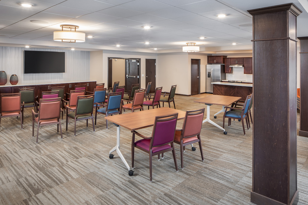 A modern community room with colorful chairs, tables, a large TV, and a kitchen area in the back.