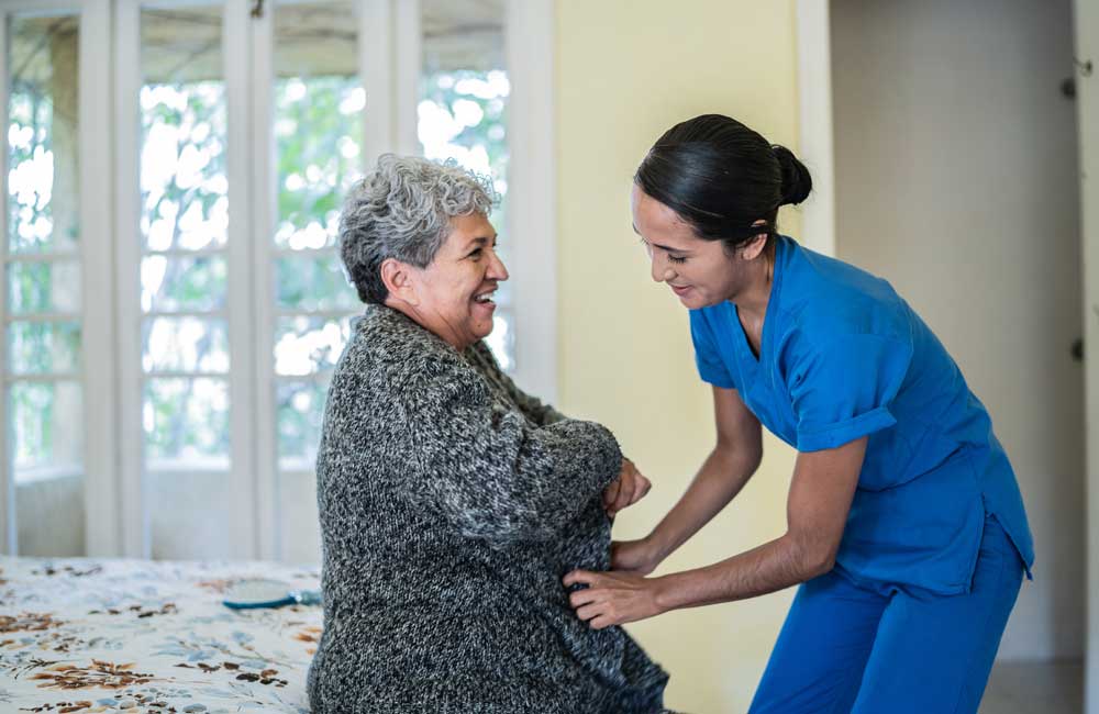 senior-grooming A smiling elderly person receives assistance from a caregiver in blue scrubs inside a bright room.