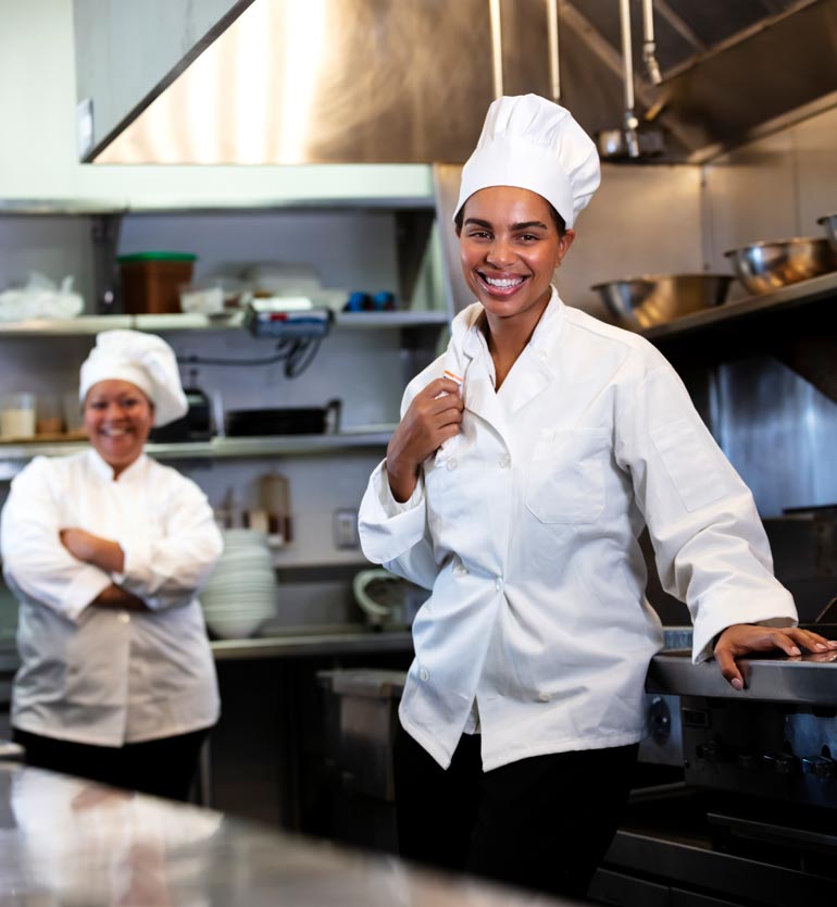 Two chefs in a professional kitchen, one smiling at the camera and one standing in the background.