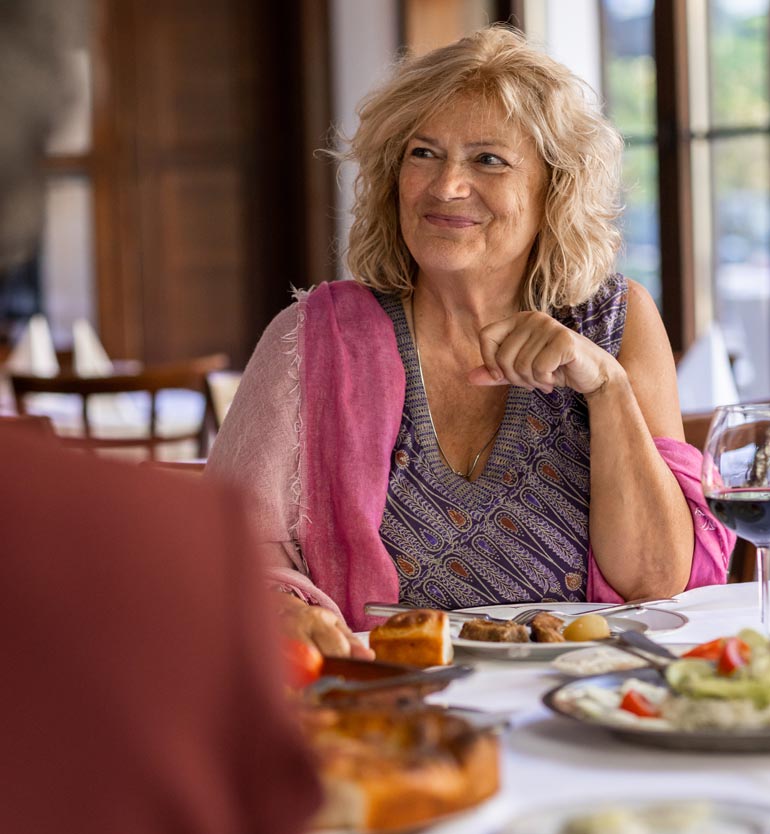 senior-friends-eating Senior woman enjoying a meal with friends at a restaurant, smiling and wearing a pink shawl.
