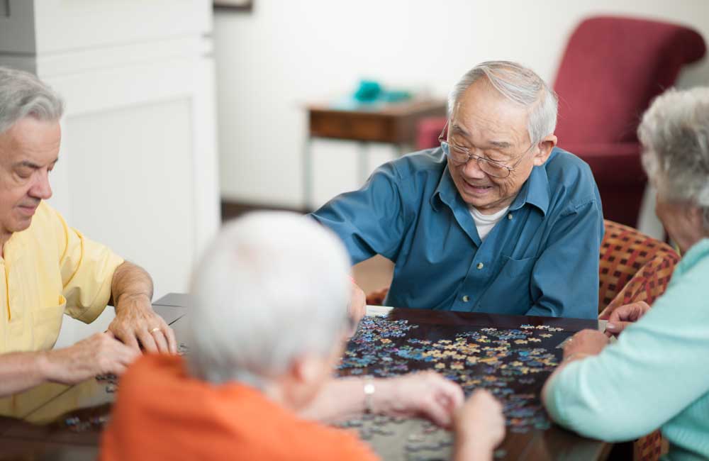 seniors-doing-puzzle Four senior individuals are enjoying putting together a jigsaw puzzle on a table at home.