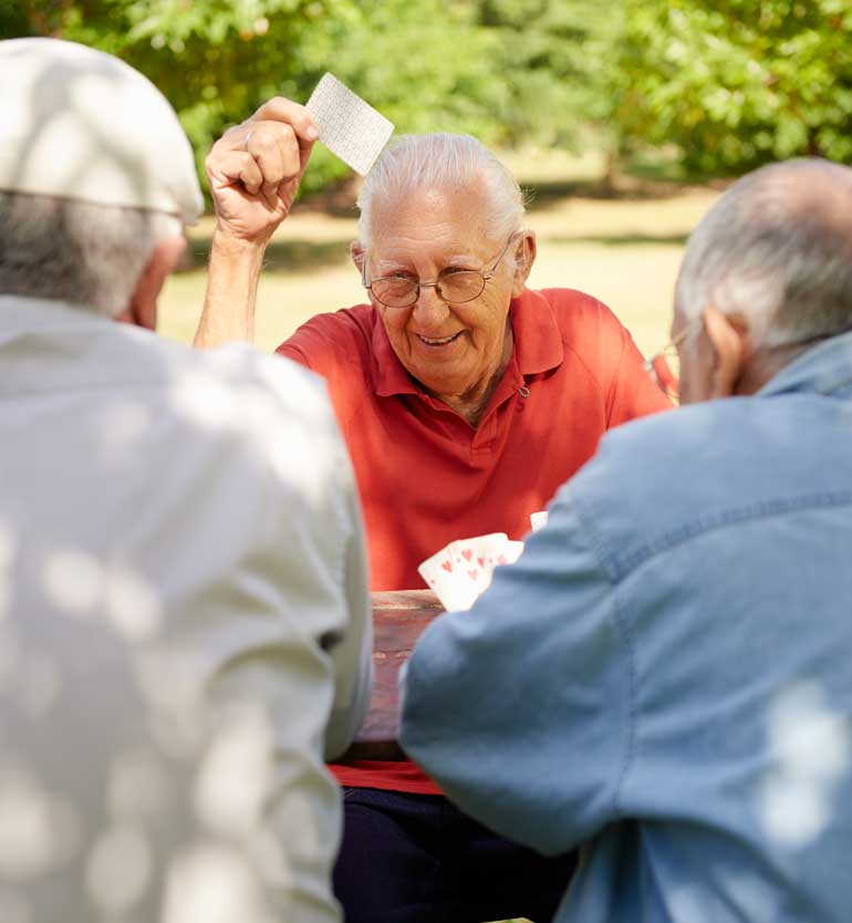 seniors-playing-cards Three seniors enjoy a lively game of cards outdoors on a sunny day at a wooden table.