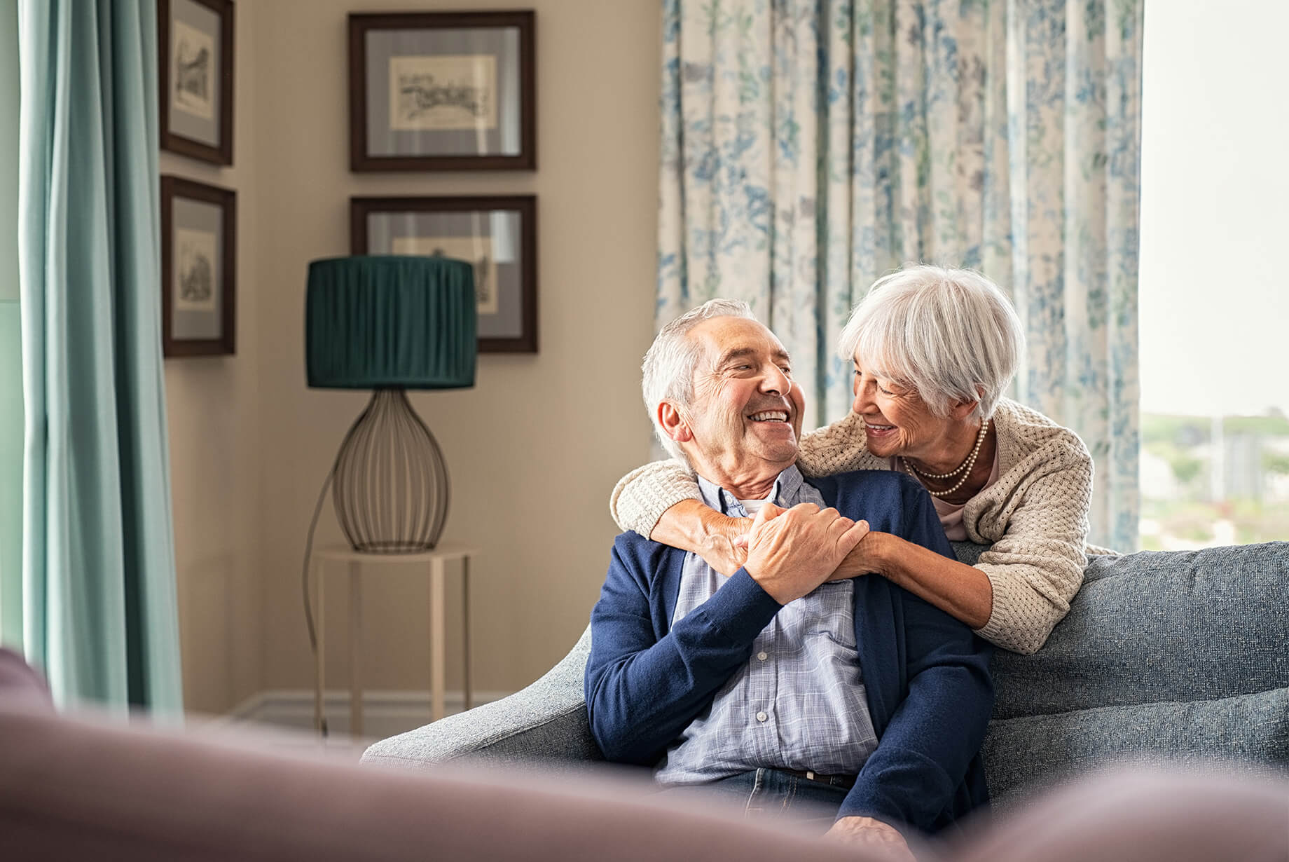 Senior woman stands behind couch and embraces senior man while they smile at each other
